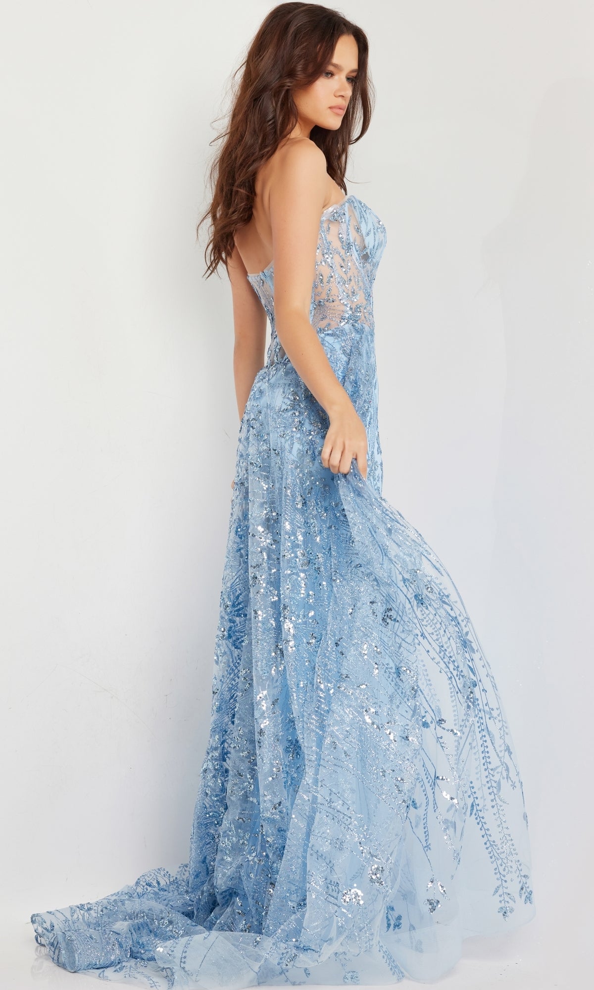 A woman wears the Jovani Strapless Long Glitter-Sequin Prom Dress 26113 in light blue, featuring a mermaid skirt and sheer corset-style bodice, standing against a plain white background.